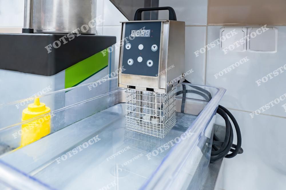 На фотографии изображено устройство для приготовления sous vide в профессиональной кухне. Аппарат для низкотемпературного приготовления расположен на рабочей поверхности, демонстрируя использование современных технологий. Снимок подчеркивает точность и инновации в ресторанном бизнесе.